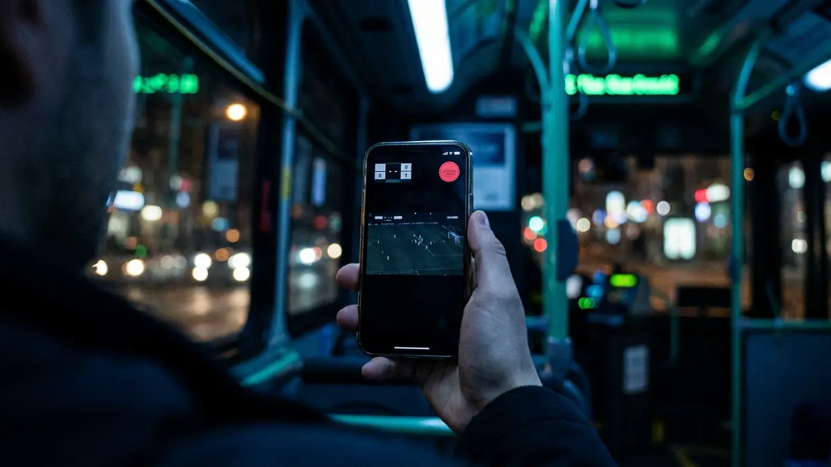 Mão segurando um celular dentro de um ônibus à noite, com a transmissão de um jogo na tela e as luzes da cidade desfocadas ao fundo.